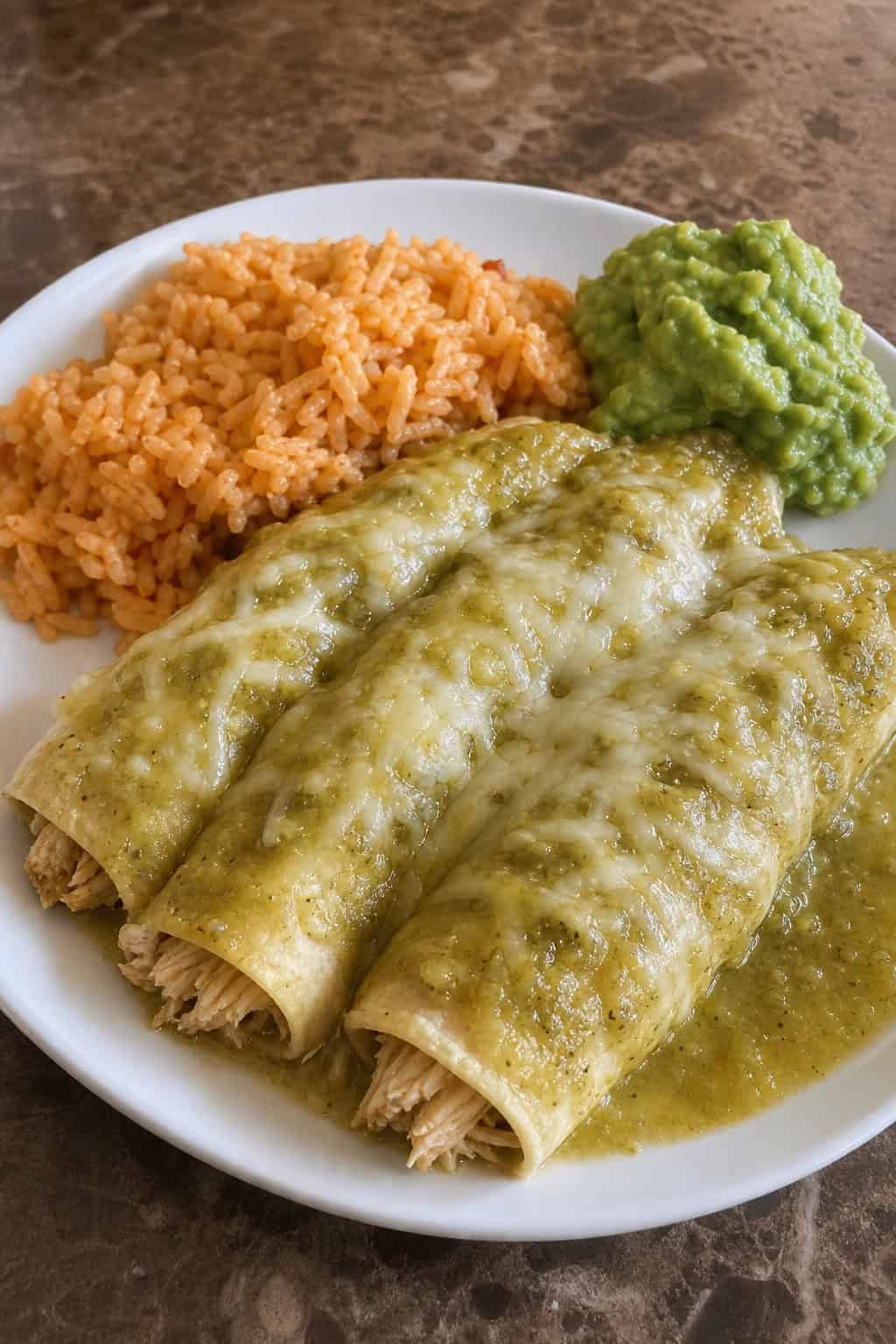 A plate of salsa verde chicken enchiladas topped with melted cheese and cilantro, served with rice and guacamole.