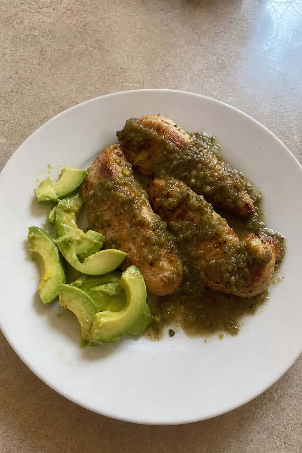 A plate of crispy chicken tenders topped with green salsa verde sauce, served with sliced avocado and garnished with cilantro and lime wedges on a wooden table.