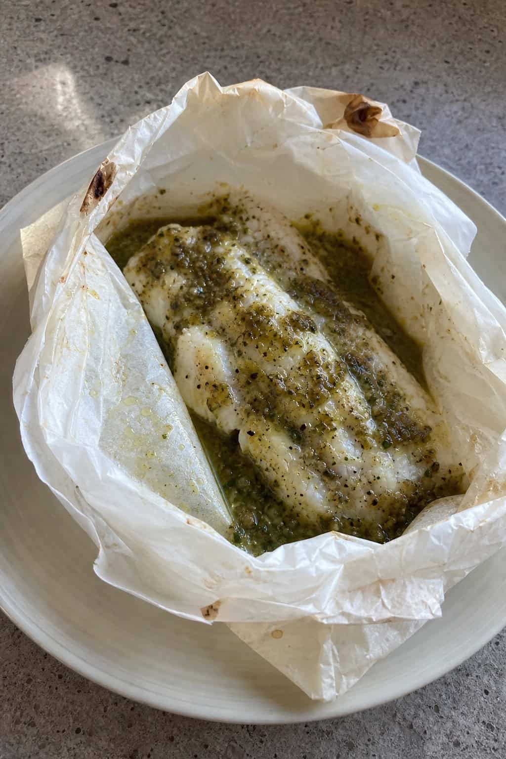 A plated fish dish wrapped in parchment paper with green salsa and garnished with fresh herbs, surrounded by tomatillo husks on a kitchen table.