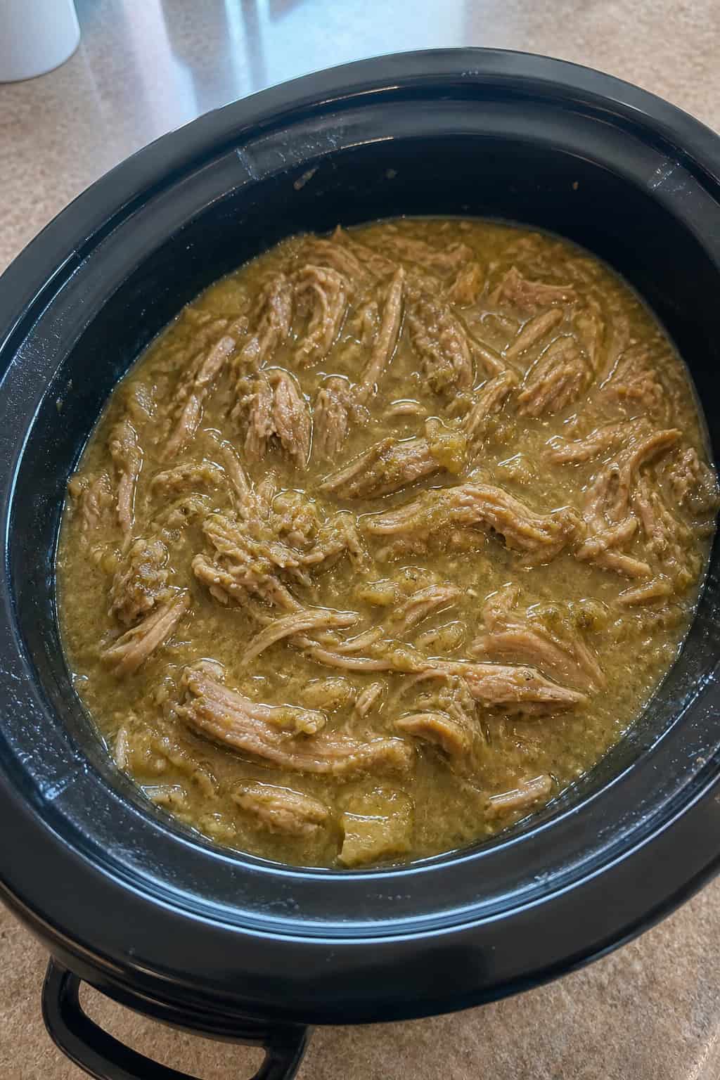 A crockpot filled with shredded pork shoulder in green salsa verde sauce, garnished with cilantro, on a wooden kitchen counter with fresh ingredients nearby.