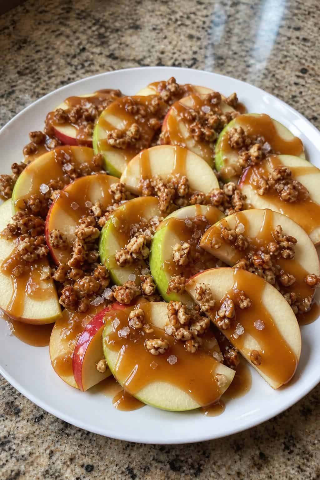A plate of thinly sliced green and red apples drizzled with salted caramel sauce and sprinkled with nuts on a wooden board.