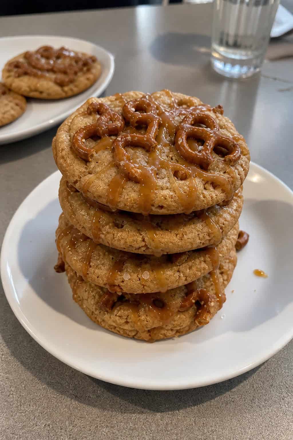 A stack of salted caramel pretzel cookies on a wooden surface with pretzels and caramel sauce nearby.
