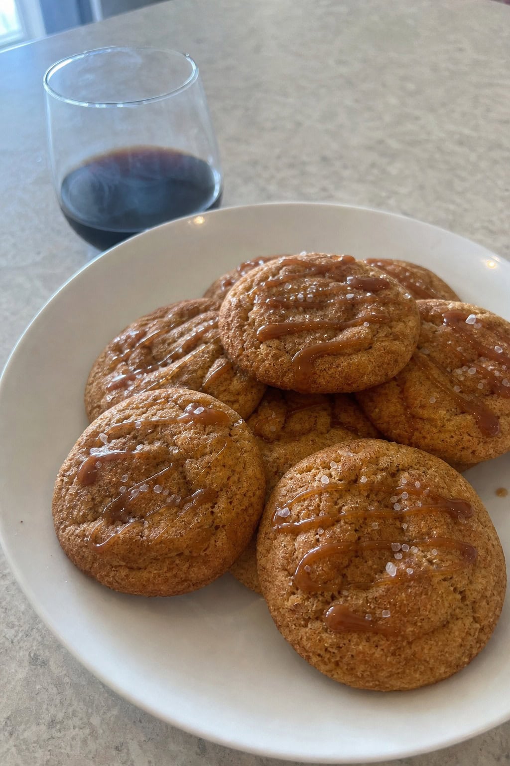 A plate of golden-brown salted caramel soy sauce snickerdoodle cookies with caramel drizzle and sea salt on a wooden surface, surrounded by cinnamon sticks and small bowls of soy sauce and caramel.