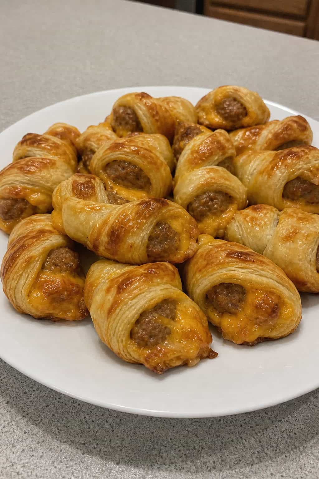 A wooden board with golden sausage and cheddar puff pastry twists, a bowl of dipping sauce, and fresh herbs on a table.