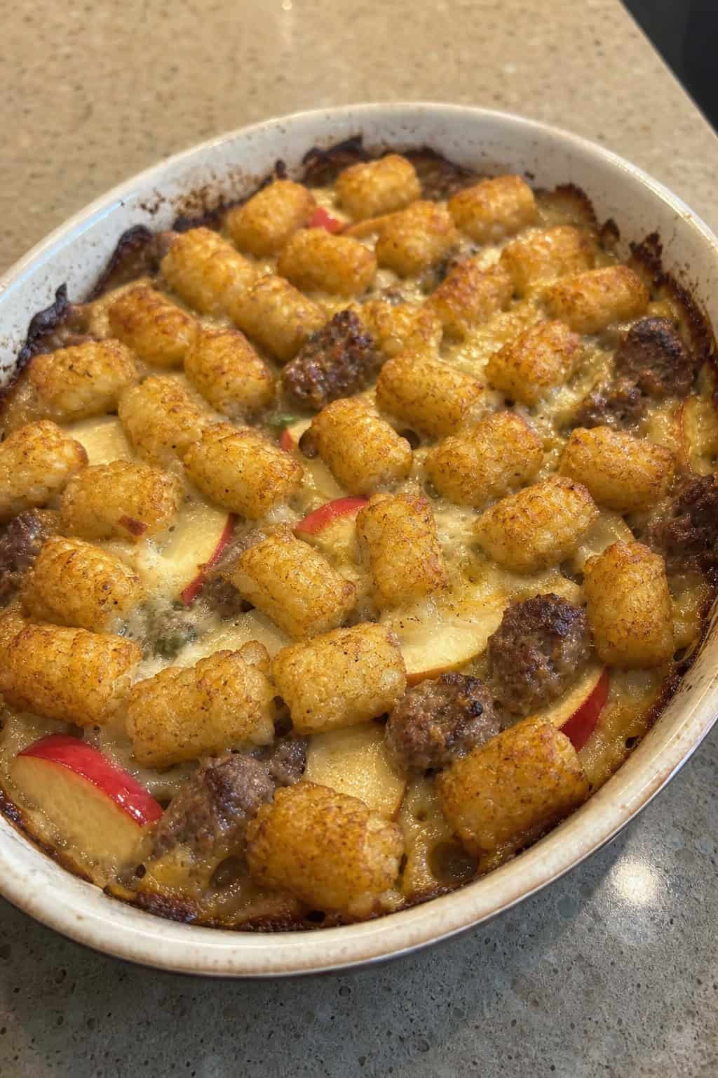 A baked casserole with crispy tater tots arranged in rows, visible sausage pieces, pale diced apple, and small green sage leaves in a cream-colored ceramic dish on a wooden table.
