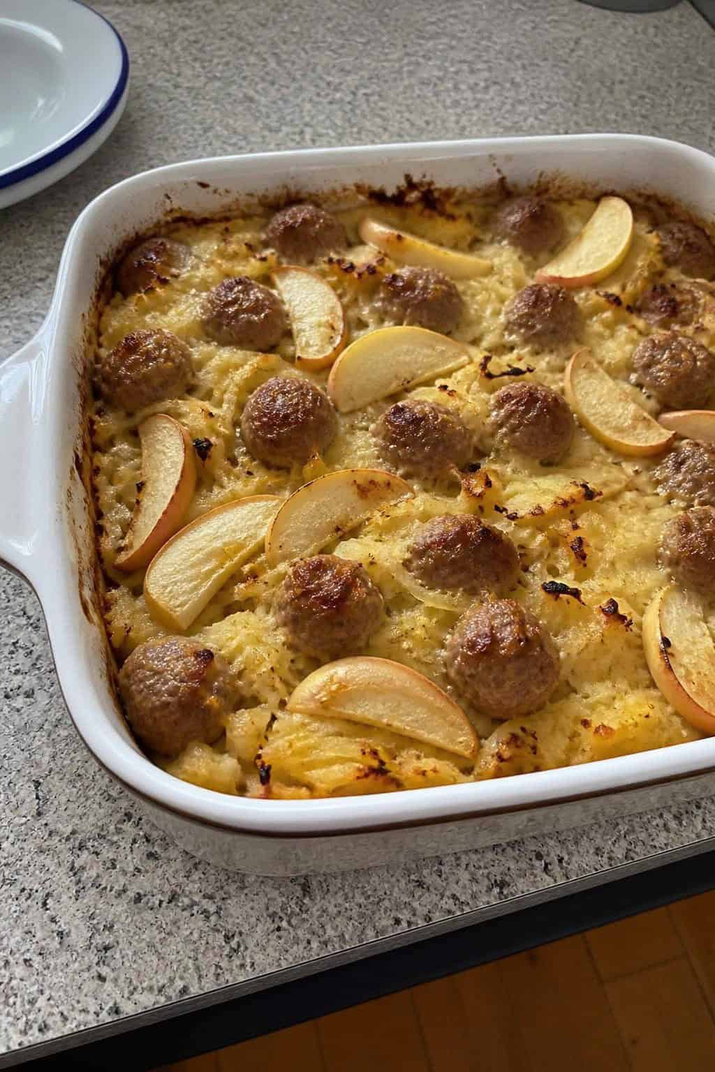 A freshly baked sausage, apple, and hashbrown casserole in a rustic baking dish on a wooden table with fresh apples and herbs nearby.