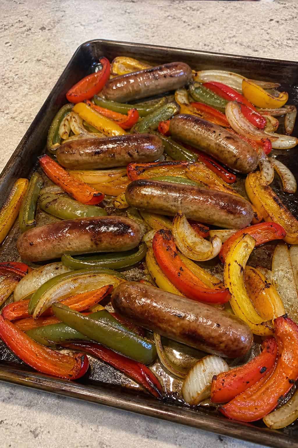 Sliced smoked sausage rounds roasted alongside strips of red, yellow, and green bell peppers and caramelized onion rings on a foil-lined sheet pan resting on a wooden table.