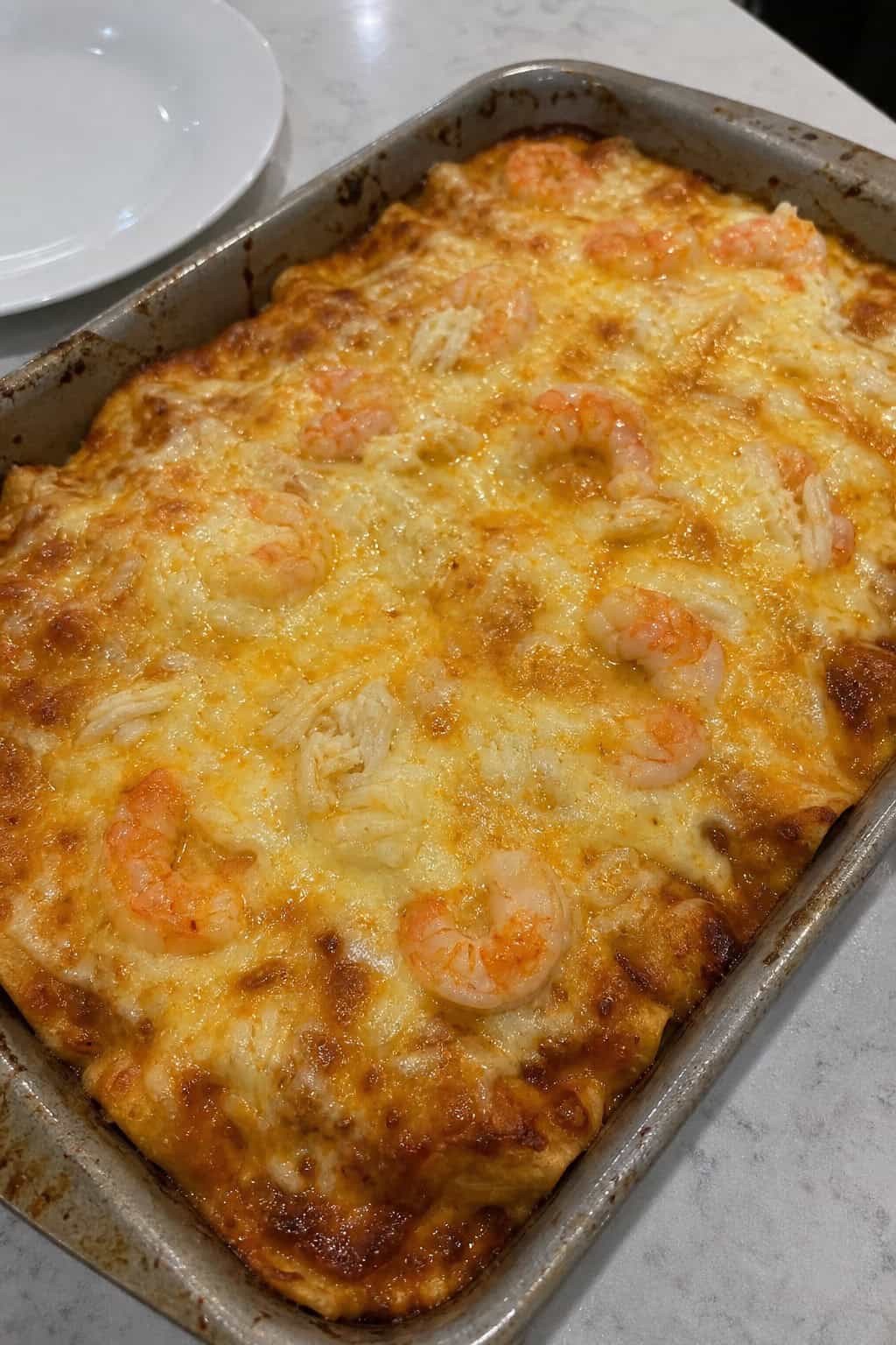 A baked seafood enchilada casserole with visible shrimp and crab in a cream sauce, garnished with fresh cilantro and lime wedges in a white ceramic baking dish on a wooden table.