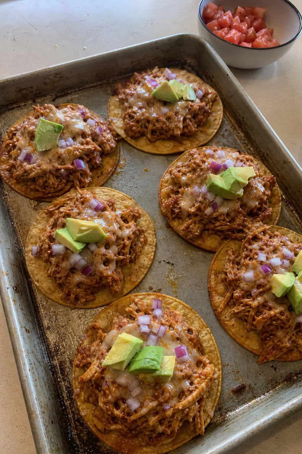 Sheet pan filled with BBQ canned chicken tacos topped with cheese, cilantro, and avocado slices on a wooden table with small bowls of salsa and lime wedges nearby.