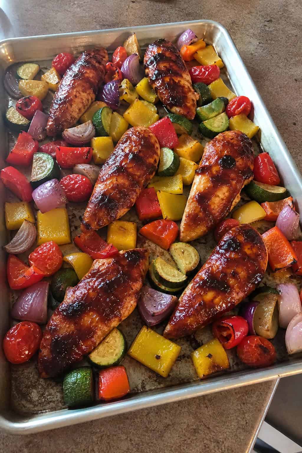 A foil-lined sheet pan with saucy BBQ chicken tenderloins on one side and roasted broccoli, red bell pepper, and baby carrots on the other.