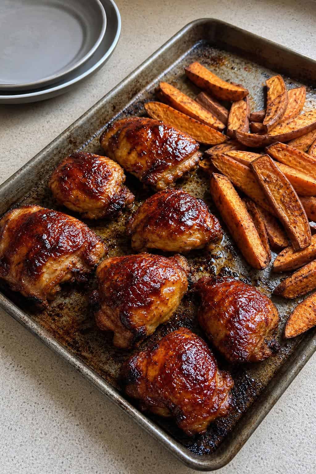 23 Healthy Chicken and Vegetable Recipes 14 A sheet pan with BBQ chicken and golden sweet potato wedges on a wooden table, surrounded by fresh vegetables and dipping sauces.