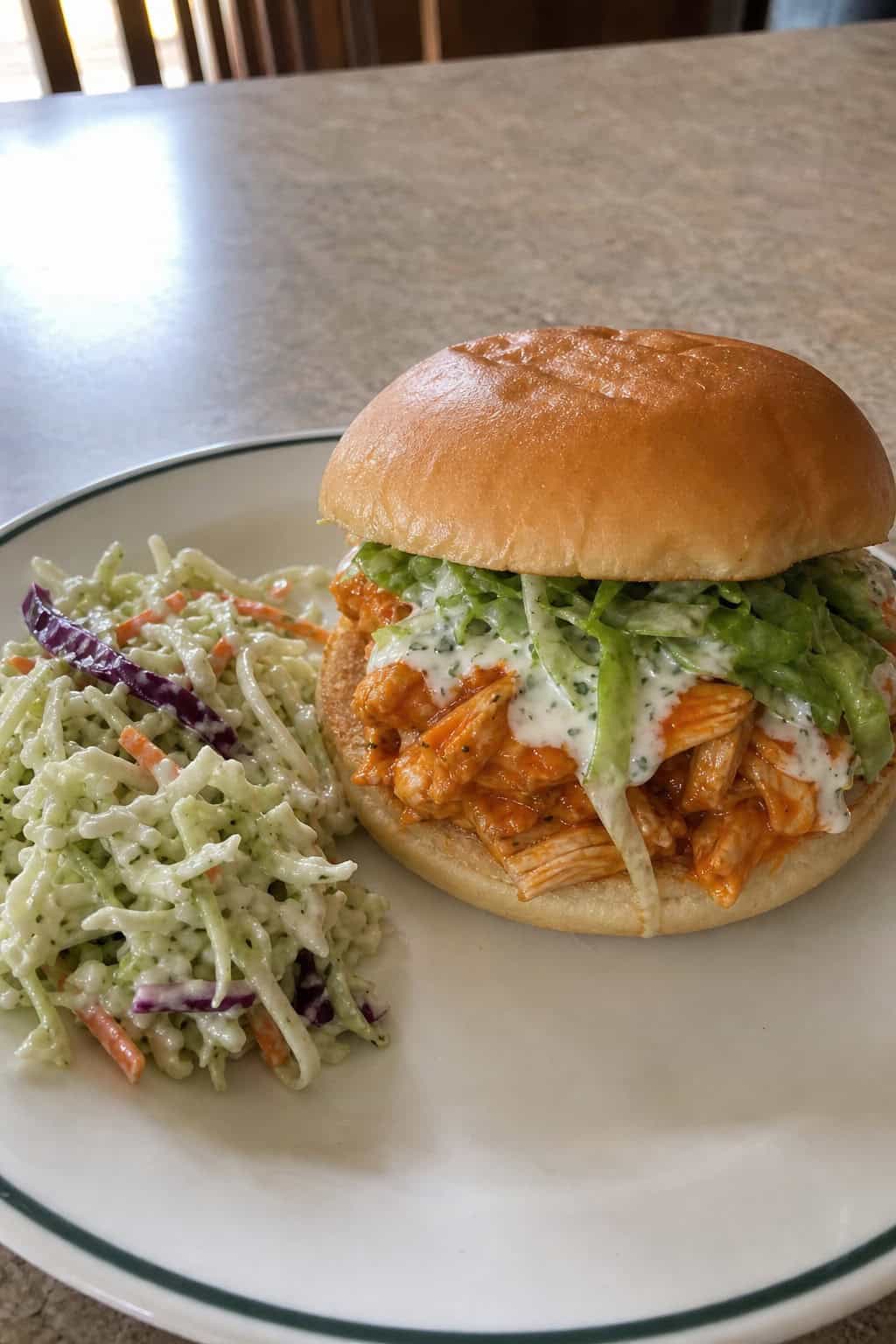 A buffalo chicken sandwich on a toasted bun with creamy ranch slaw spilling out the sides, served next to a bowl of extra slaw