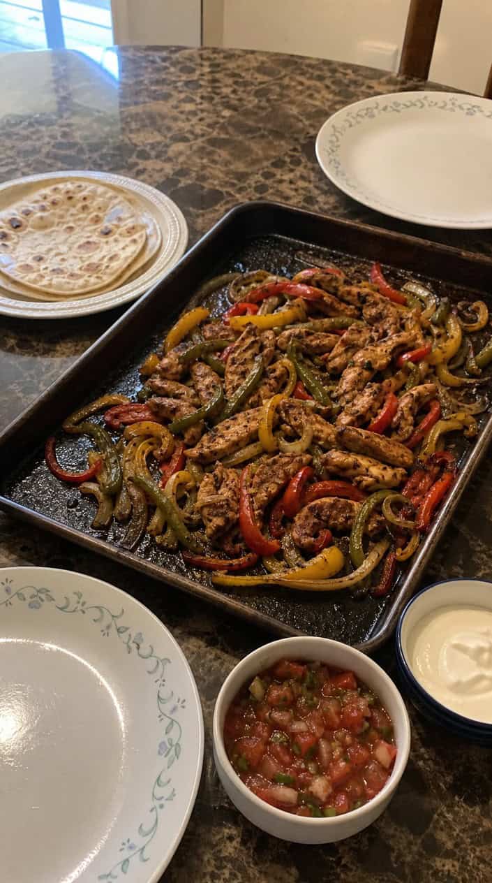 A sheet pan filled with cooked chicken fajitas including grilled chicken strips, bell peppers, and onions, with tortillas and lime wedges on the side.