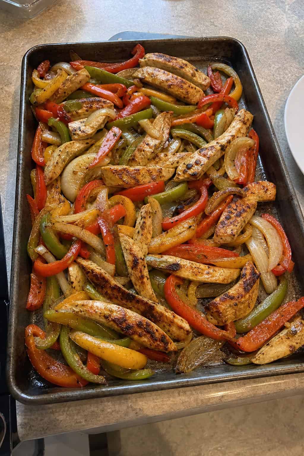 A foil-lined sheet pan filled with roasted chicken strips, caramelized red, yellow, and green bell pepper slices, and softened onion wedges, with lime wedges and a small bowl of salsa on the wooden table nearby.
