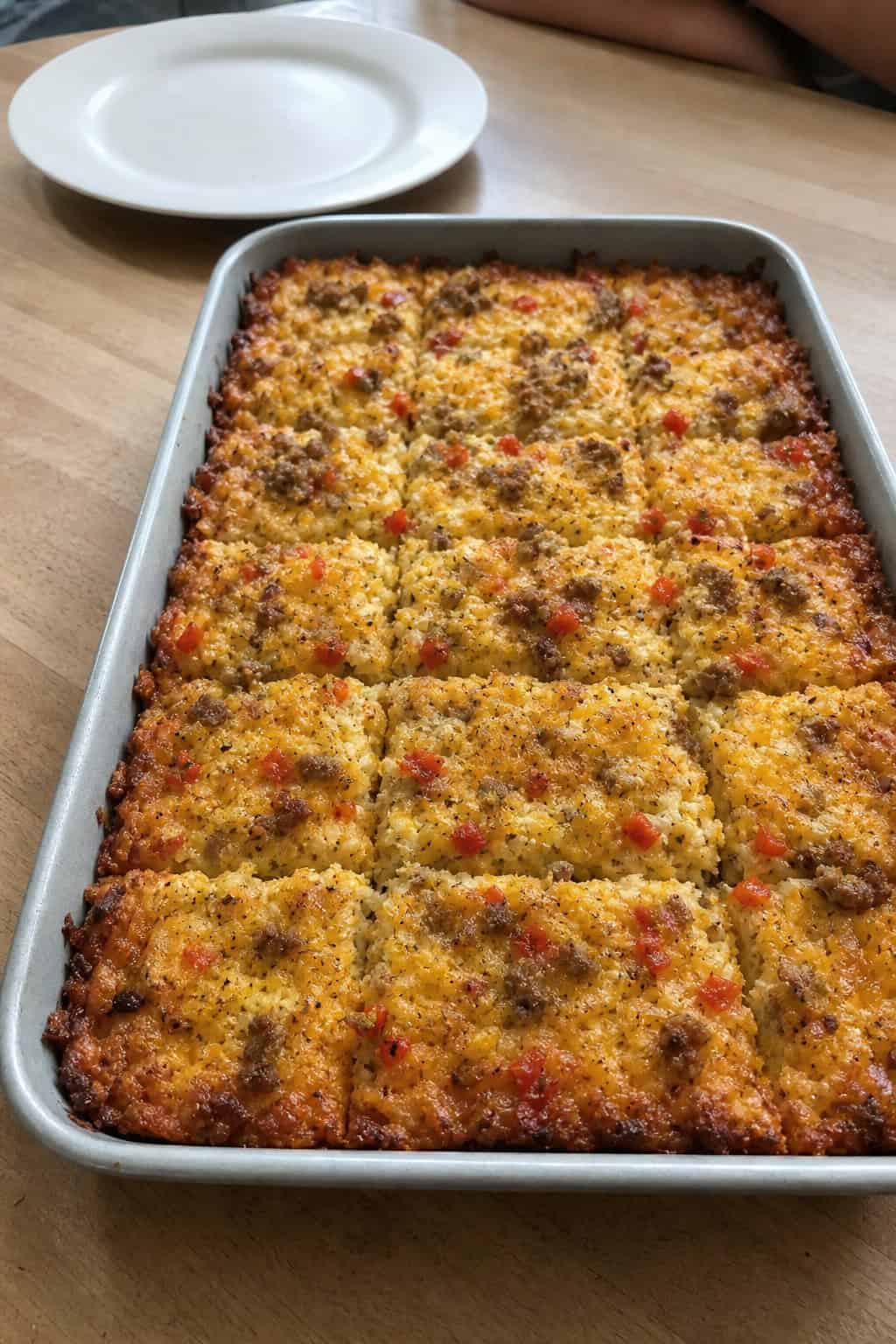 A sheet-pan hashbrown casserole cut into neat squares on a wood table, with golden-brown crispy edges and a cheesy top, surrounded by a few fresh ingredients.