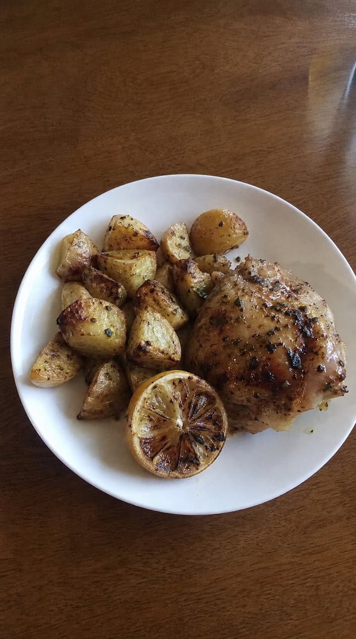 Sheet-pan lemon herb chicken and roasted potatoes on a wooden table outdoors with a backyard grill in the background.