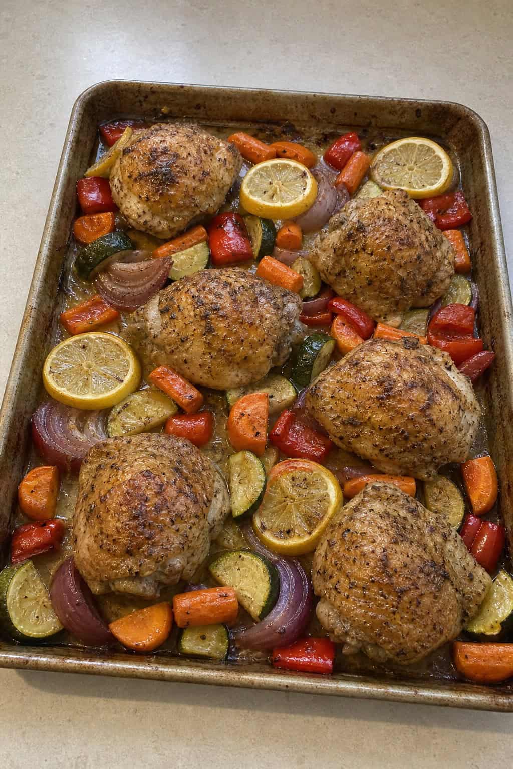 A foil-lined sheet pan loaded with lemon herb chicken thighs surrounded by roasted carrots, zucchini slices, and bright red bell pepper strips.