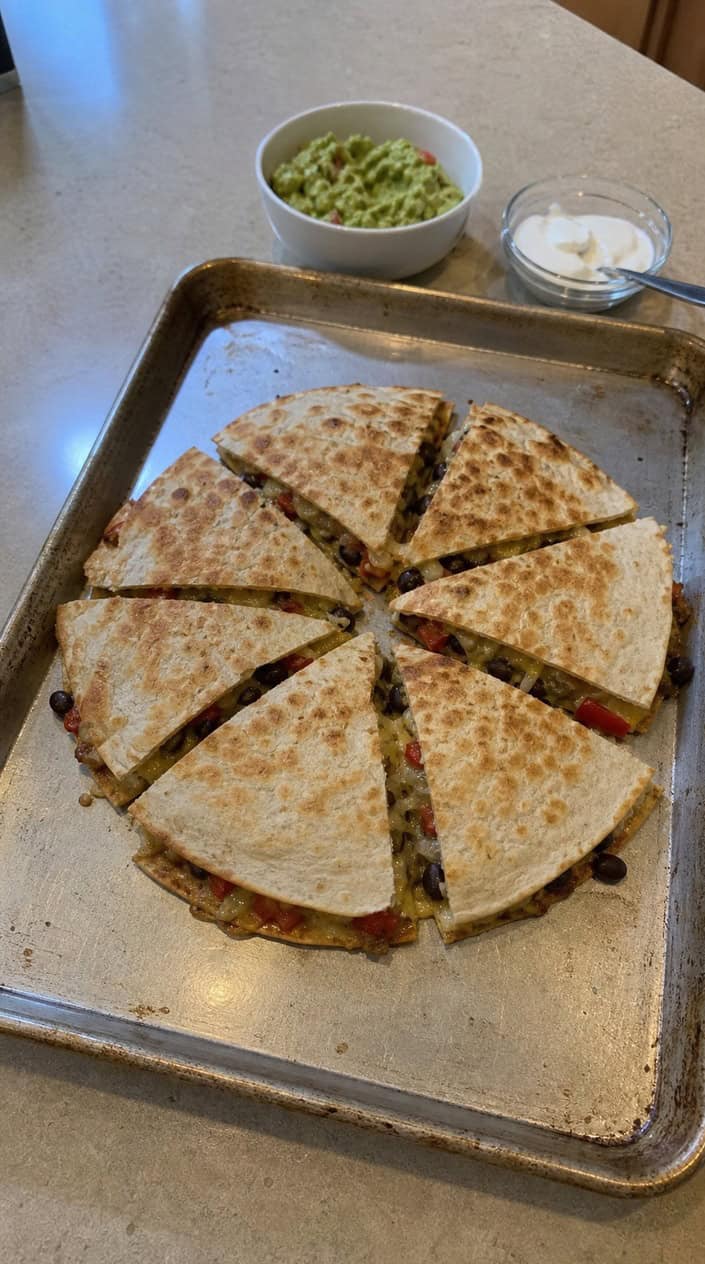 A golden-edged sheet pan quesadilla cut into triangular wedges on a kitchen countertop, revealing melted cheese and visible corn and red onion, with small bowls of salsa and sour cream alongside.