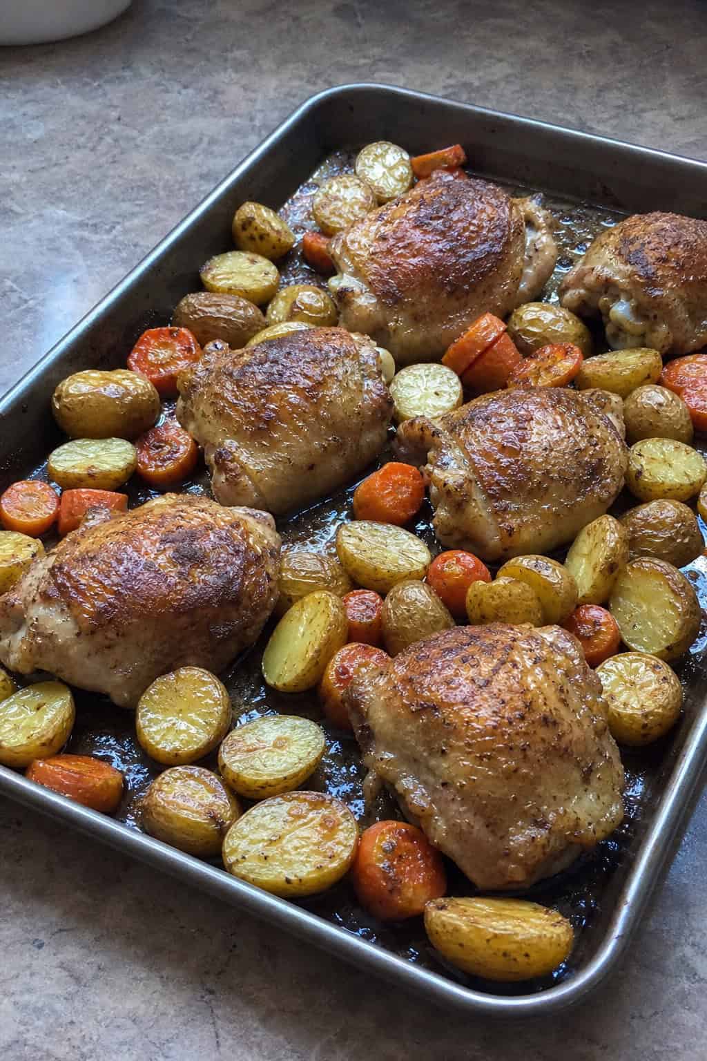 Golden roasted chicken thighs with crispy skin resting on a bed of halved baby potatoes and carrot sticks on a well-seasoned rimmed metal baking sheet.