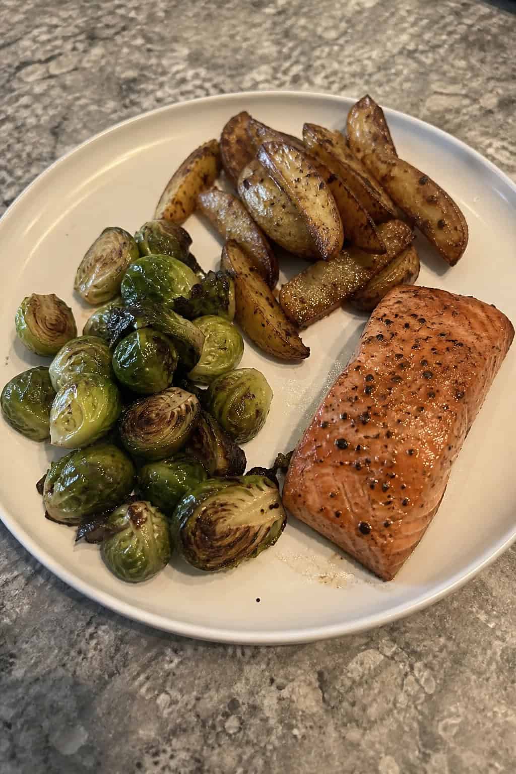 A sheet-pan meal with roasted salmon, Brussels sprouts, and potato wedges on a wooden table.
