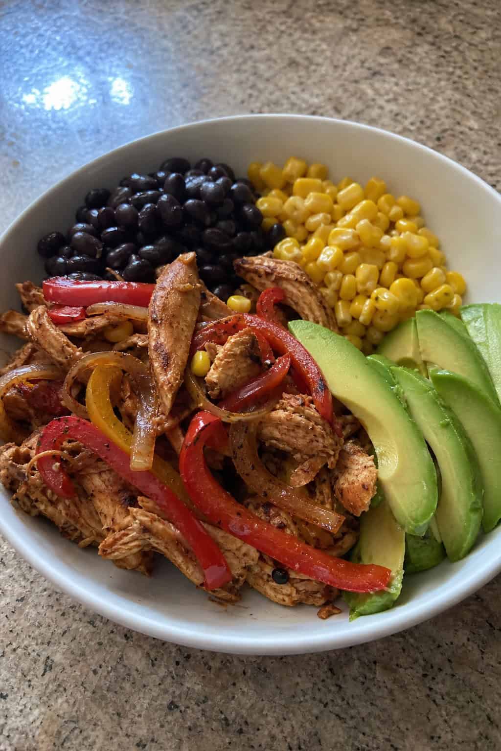 A colorful fajita bowl on a wooden table with strips of roasted red and yellow bell pepper, caramelized onion, shredded spiced chicken, sliced avocado, black beans, corn, and a lime wedge.