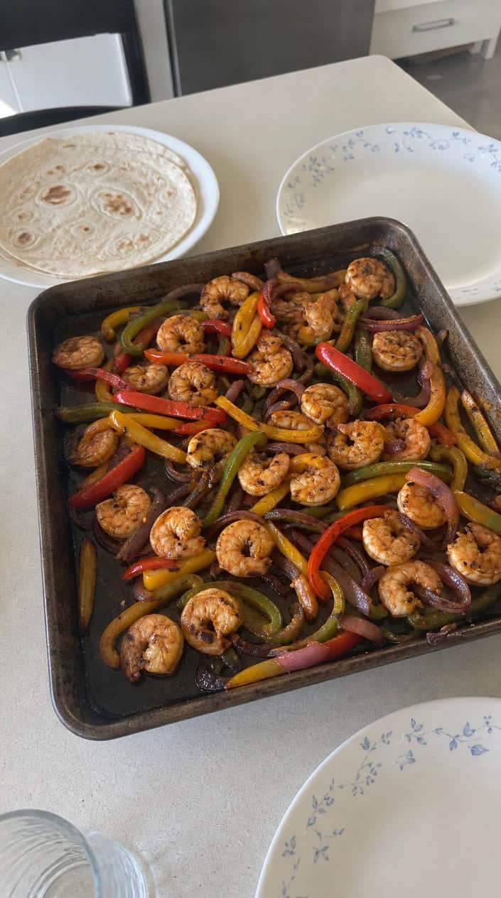 A sheet pan filled with cooked shrimp, colorful bell peppers, and onions on a wooden table with bowls of lime wedges, cilantro, and jalapeños nearby.