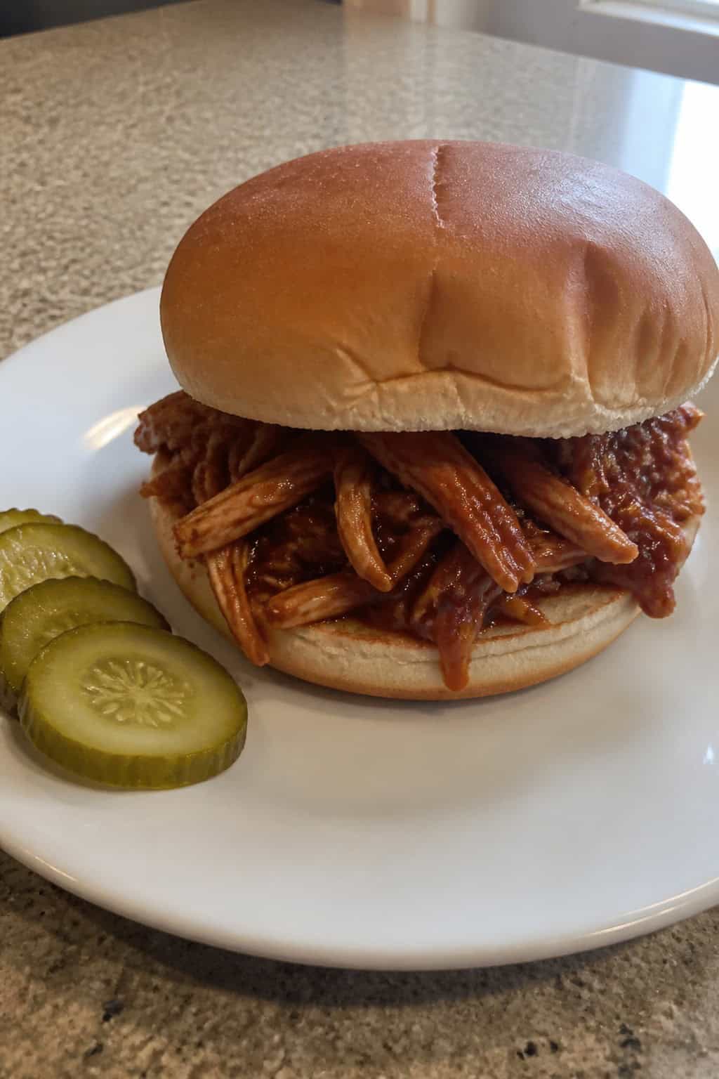 A shredded BBQ chicken sandwich on a toasted bun with lettuce and pickles on a wooden board.