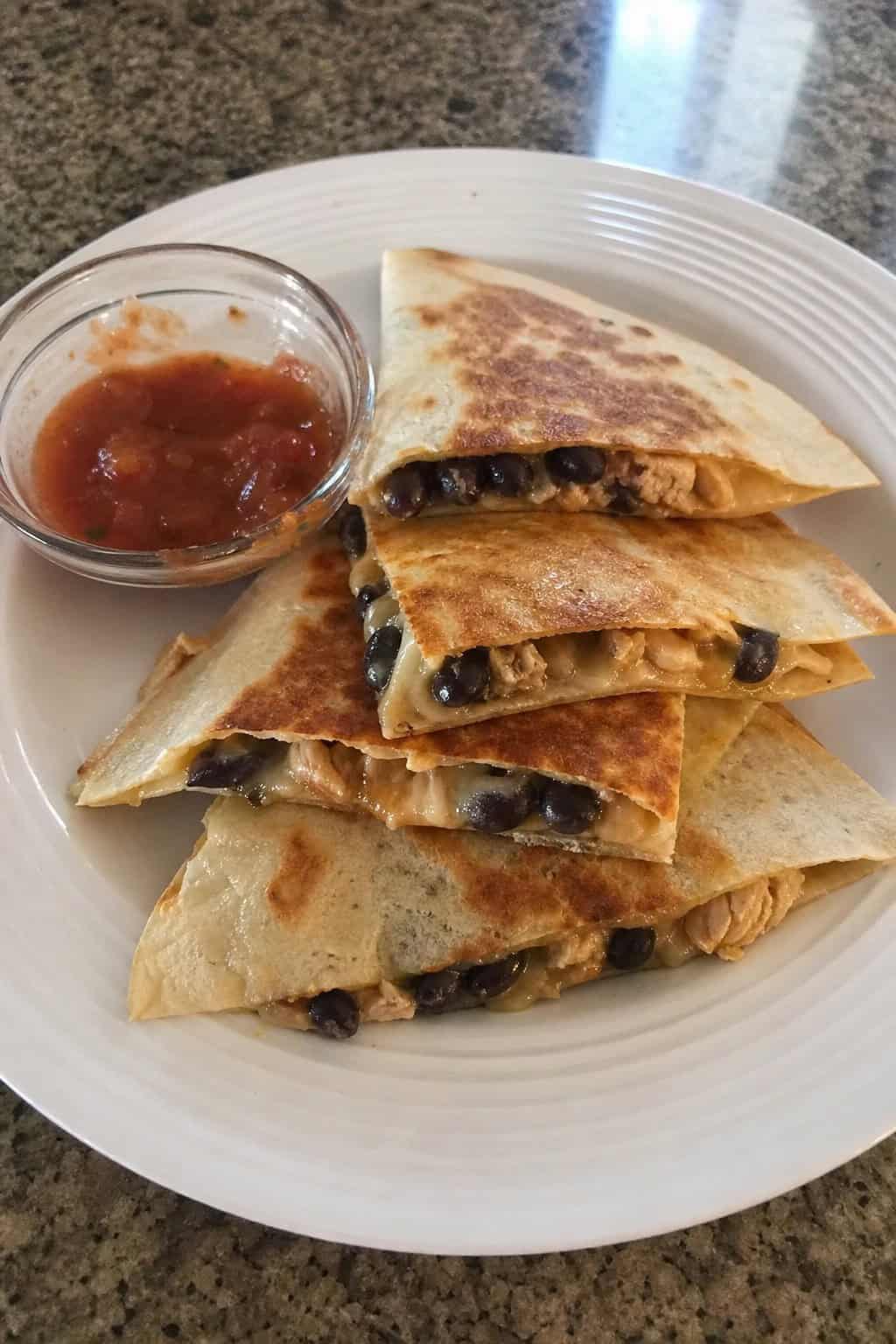 A wooden cutting board with golden-brown folded quesadillas sliced into wedges, showing a melted cheese and shredded chicken filling, with small bowls of salsa, sour cream, and lime wedges nearby.