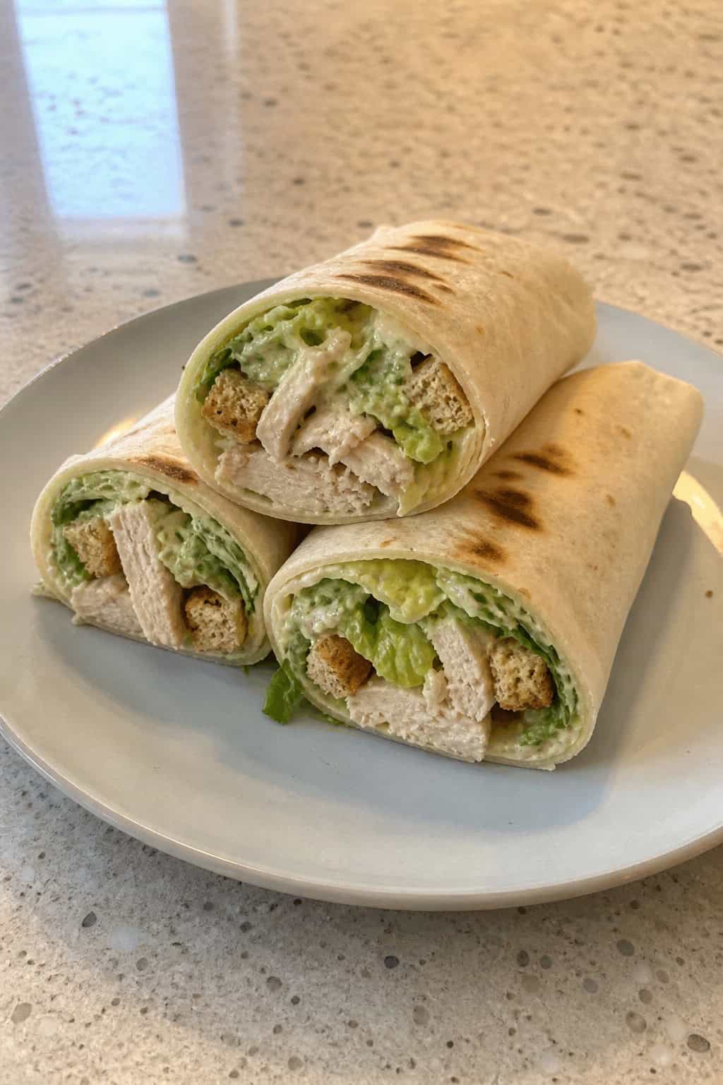 A wooden board with two shredded chicken Caesar salad wraps sliced on the diagonal, showing layers of romaine, Parmesan, and creamy dressing, with lemon wedges and a small pile of extra croutons alongside.