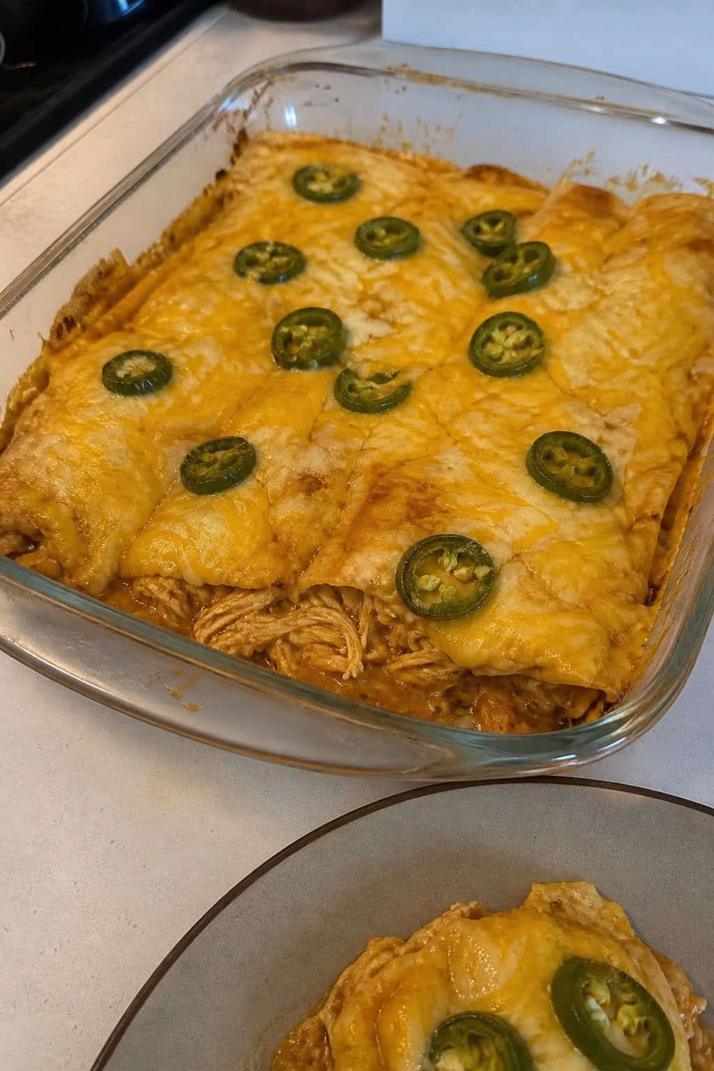 A rectangular baking dish of shredded chicken enchilada casserole fresh from the oven, with bubbly melted cheese on top and thin slices of jalapeño and a scattering of fresh cilantro leaves.