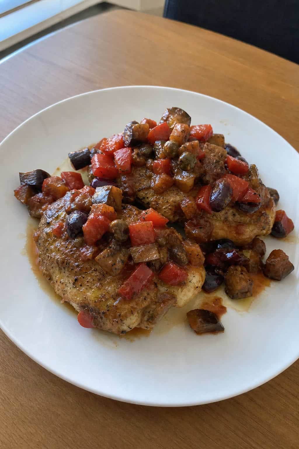 A plate of golden chicken thighs topped with colorful Sicilian caponata, placed on a wooden table with fresh herbs and a glass of red wine nearby.