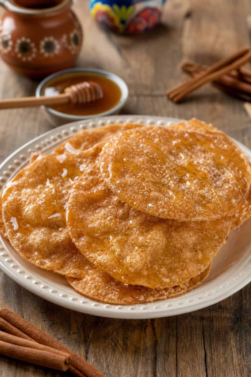 A plate of golden Mexican buñuelos drizzled with honey on a wooden table, with small bowls of honey and cinnamon sticks nearby.