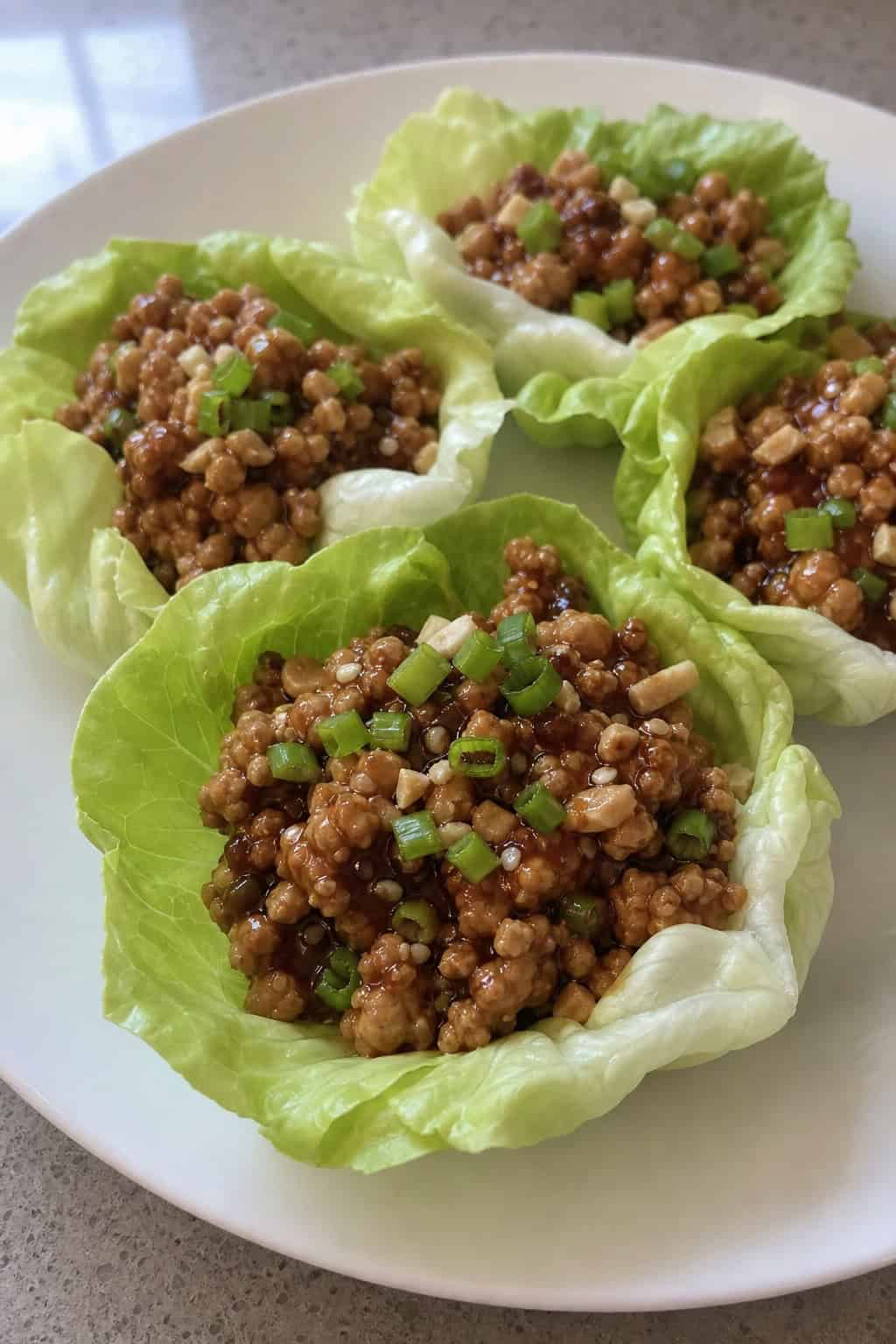 Close-up of minced chicken lettuce wraps filled with seasoned chicken and garnished with green onions and sesame seeds on a wooden platter with small bowls of sauce and herbs nearby.