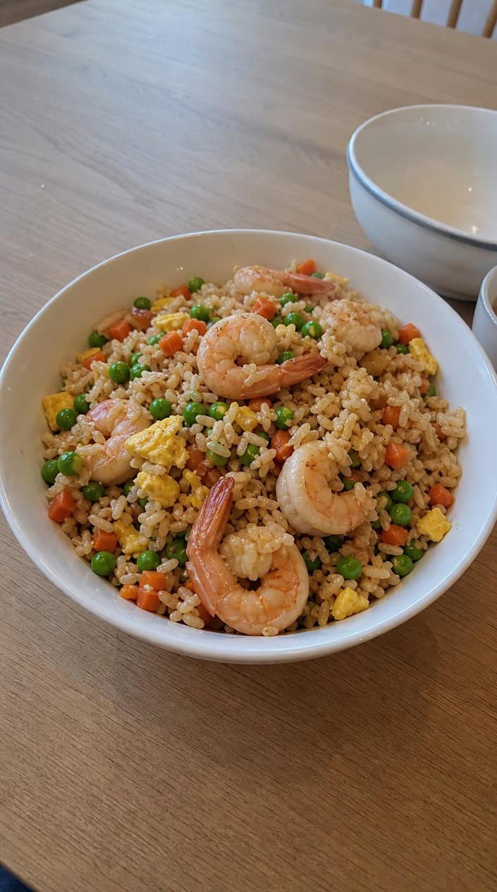 A bowl of shrimp fried rice with colorful vegetables on a wooden table surrounded by fresh ingredients.