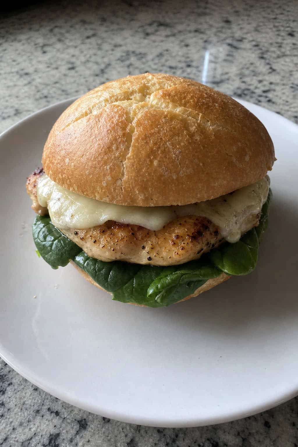 A pan-seared chicken sandwich with baby spinach on a toasted bun, set on a wooden table with a bowl of greens and a glass of iced tea nearby