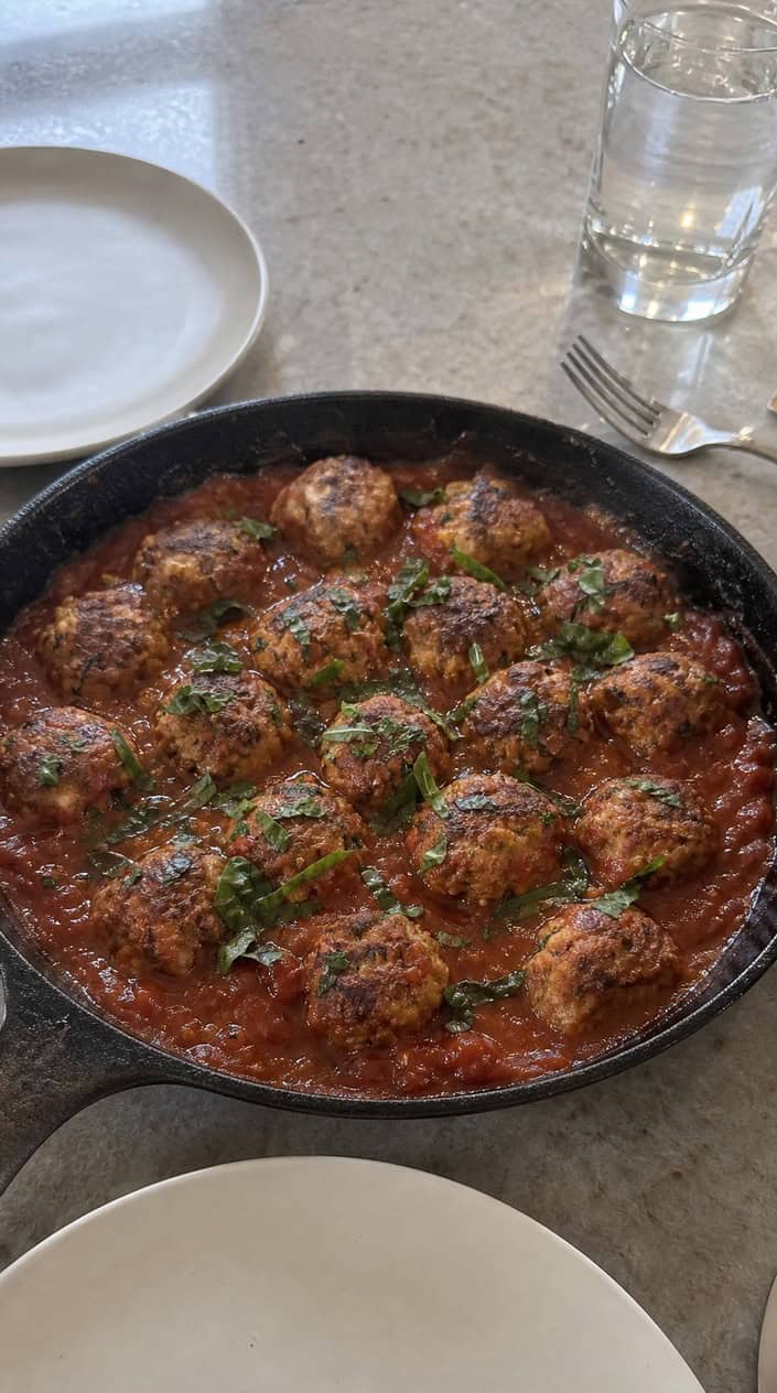 A skillet with chicken meatballs in tomato and basil sauce on a wooden surface, garnished with fresh basil leaves and cherry tomatoes.