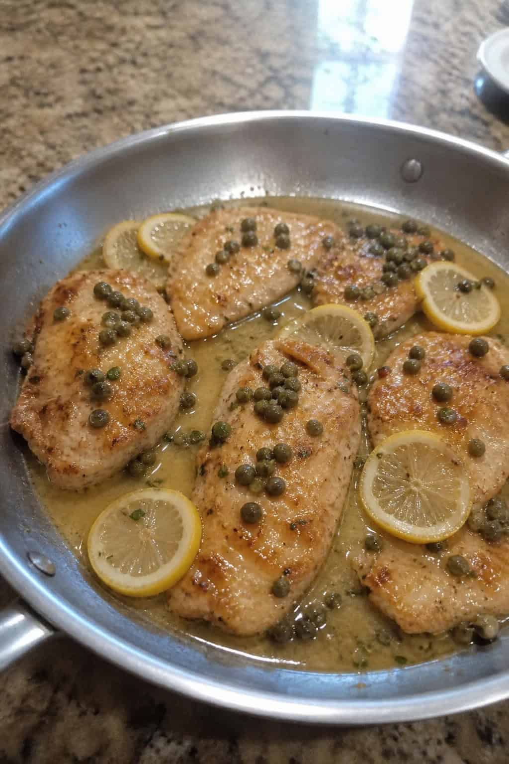 A skillet with cooked chicken piccata topped with capers and lemon slices on a wooden table with fresh ingredients around it.