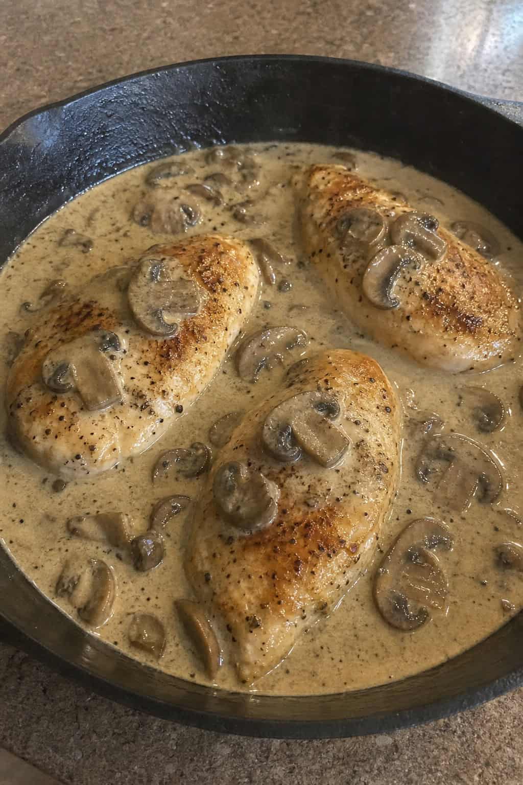 Skillet with golden chicken breast topped with creamy mushroom sauce and garnished with fresh herbs on a wooden table.