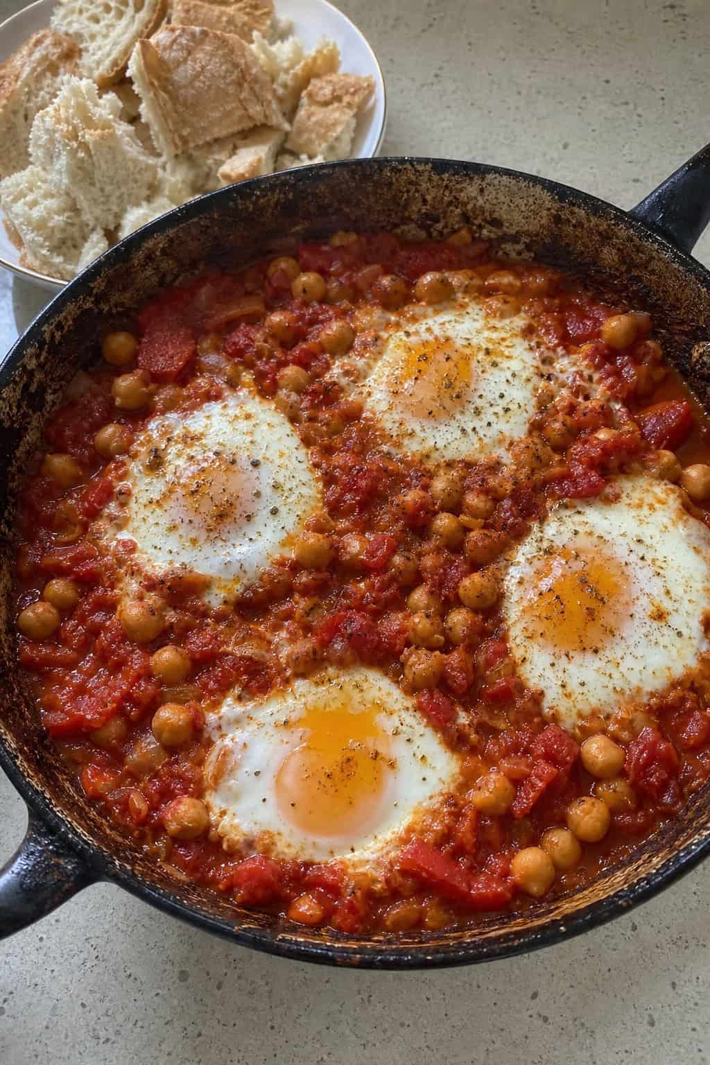 A skillet of chickpea shakshuka with poached eggs and fresh herbs, served with torn pieces of stale bread on a wooden table.