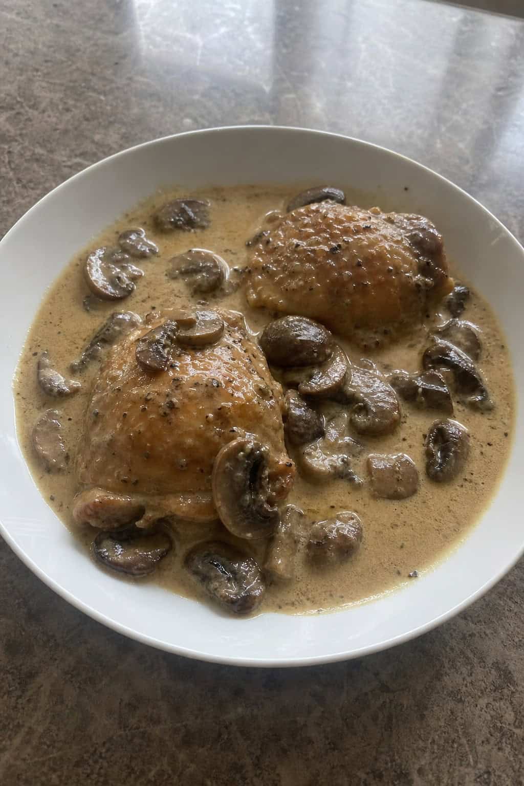 A cast iron skillet on a wooden table holding golden-seared chicken thighs nestled in a pale, creamy mushroom sauce with fresh herb sprigs alongside.