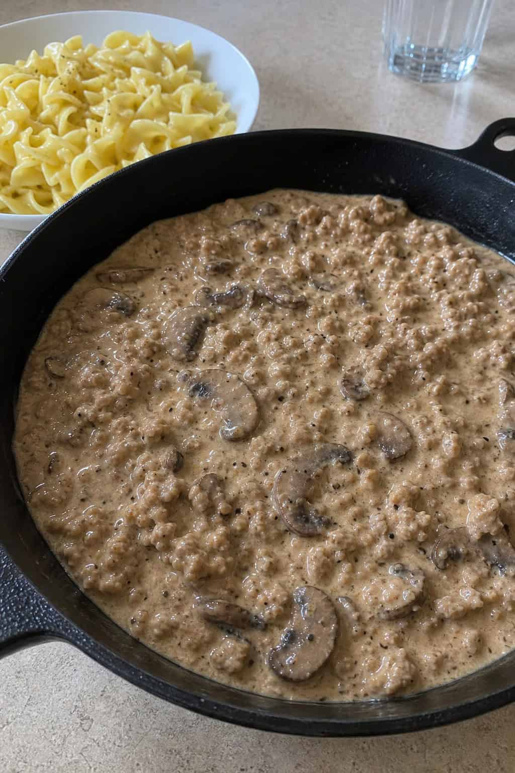 A skillet filled with creamy minced chicken and mushroom stroganoff garnished with parsley, served with a side of noodles on a wooden table.