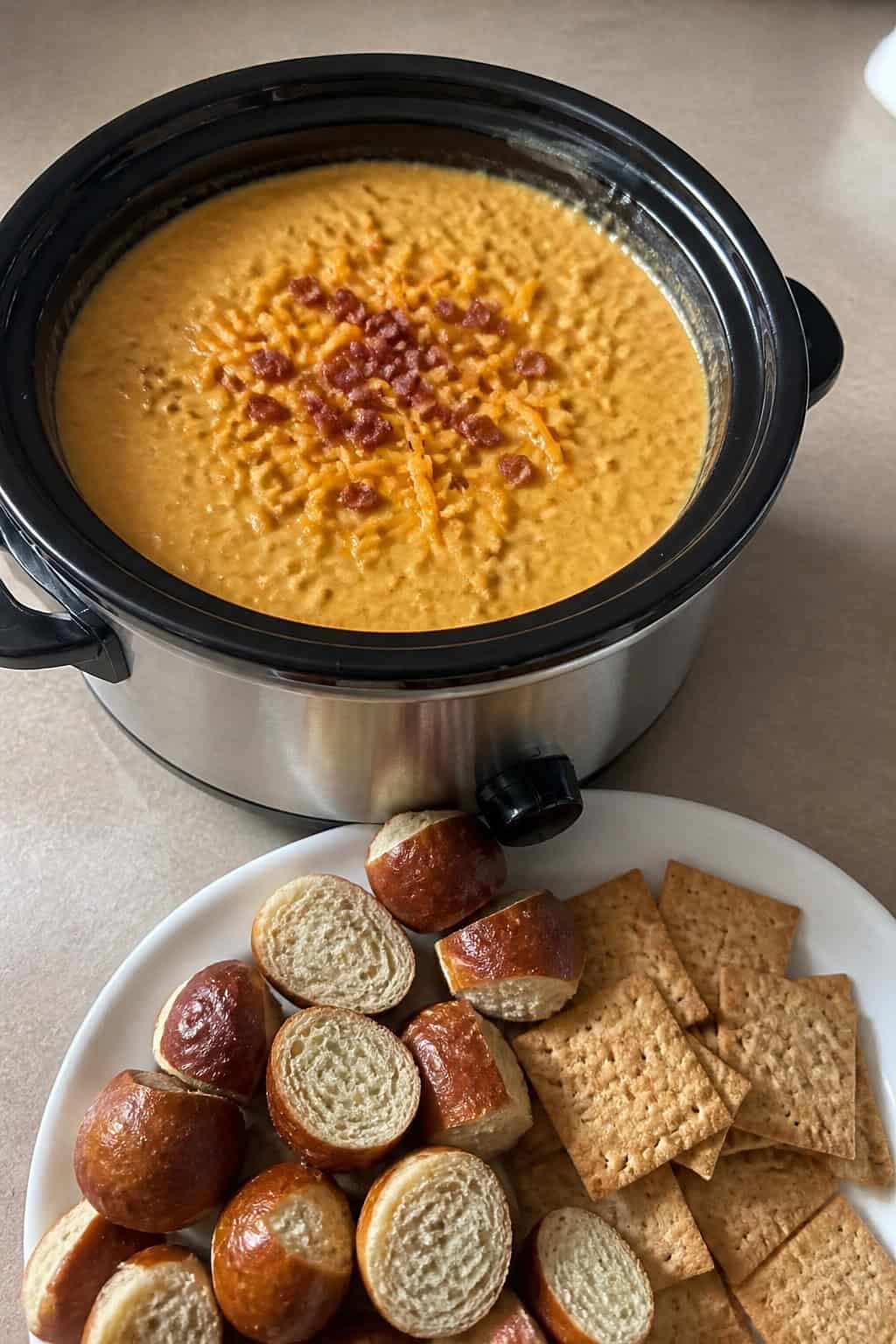 Slow cooker filled with creamy Bavarian beer cheese dip surrounded by pretzel bites, bread slices, vegetable sticks, and crackers on a wooden table with a glass of beer in the background.