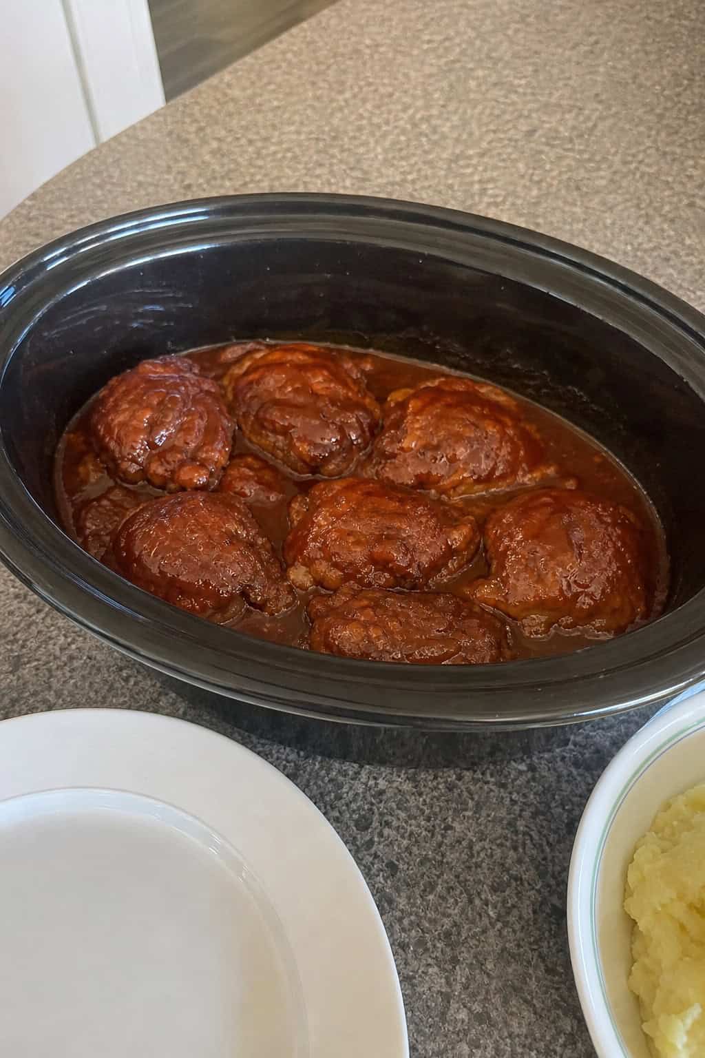 A ceramic slow-cooker insert filled with saucy BBQ chicken thighs, with fresh herbs and seasoning ingredients arranged on the countertop beside it.