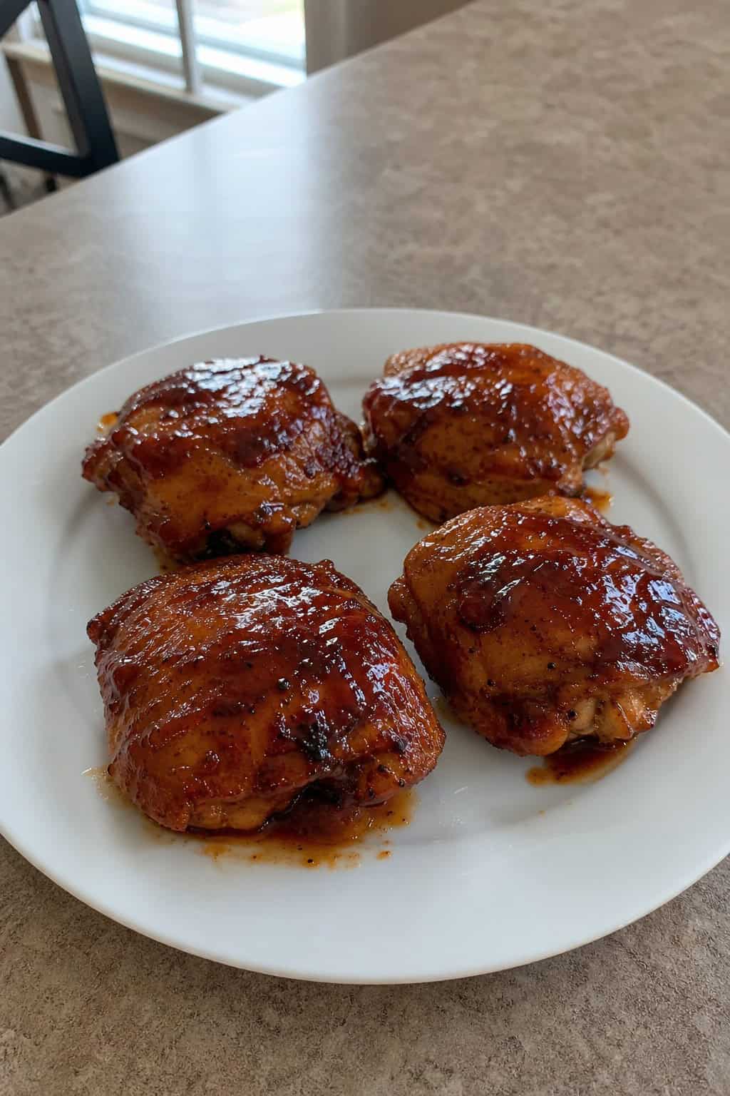 A plate of glazed BBQ bourbon chicken thighs garnished with parsley on a rustic table with a small bowl of sauce and a glass of bourbon nearby.