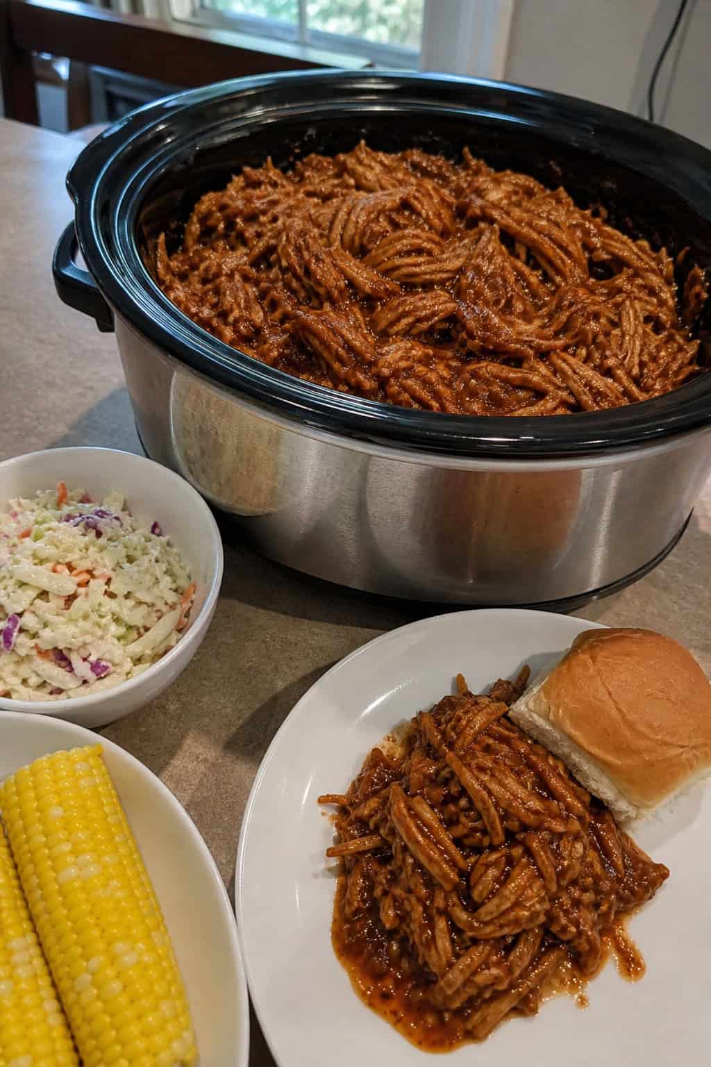 Slow-cooker filled with shredded BBQ bourbon pulled pork on a wooden table surrounded by coleslaw, corn on the cob, and a glass of iced tea.
