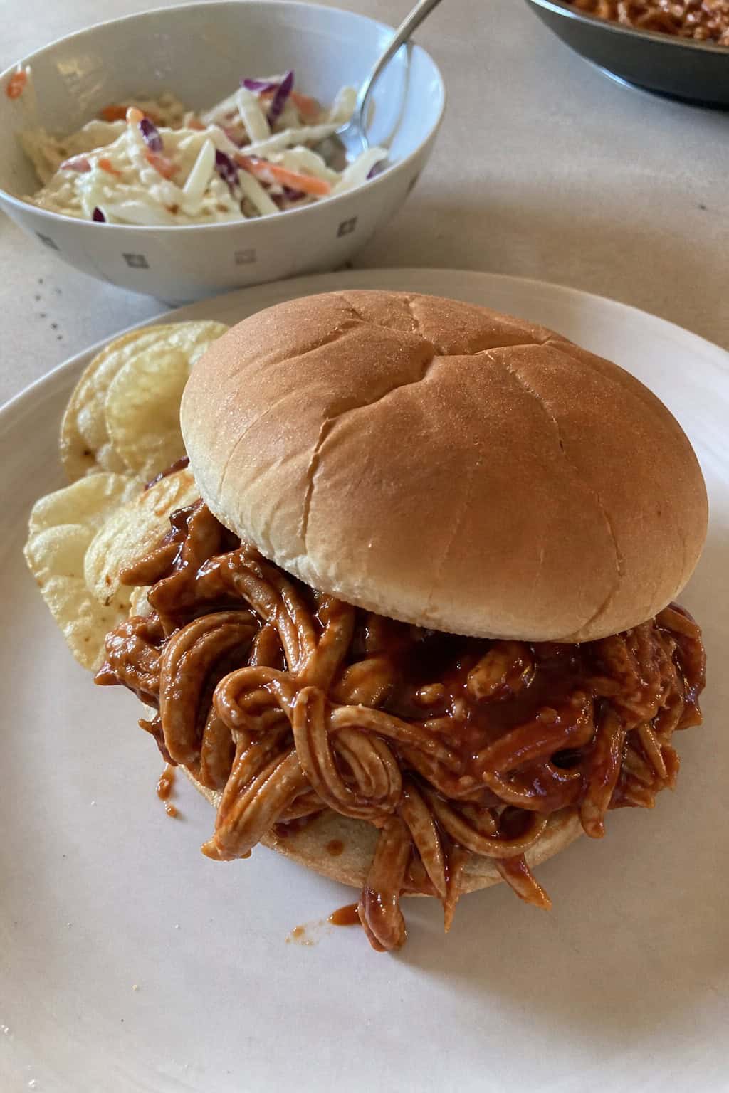 A close-up of a pulled chicken sandwich with barbecue sauce on a toasted bun, served with coleslaw and extra sauce on a wooden board.