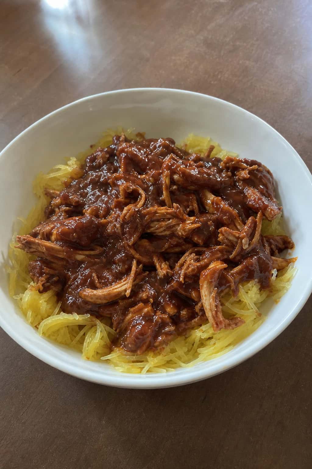 A bowl of BBQ pulled chicken served over spaghetti squash strands, garnished with fresh herbs, on a wooden table.
