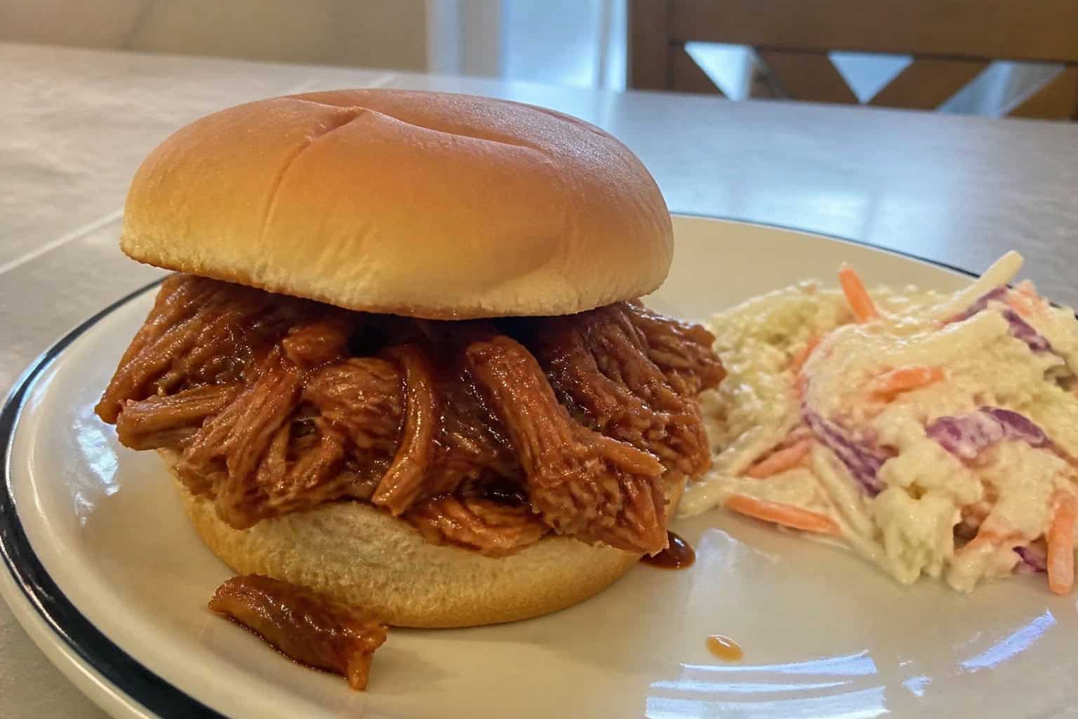 A shredded BBQ chicken sandwich on a bun with lettuce and coleslaw on a wooden table.