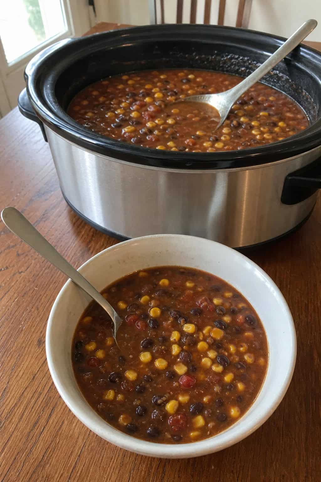 A slow cooker filled with black bean and corn soup surrounded by fresh ingredients on a kitchen counter.
