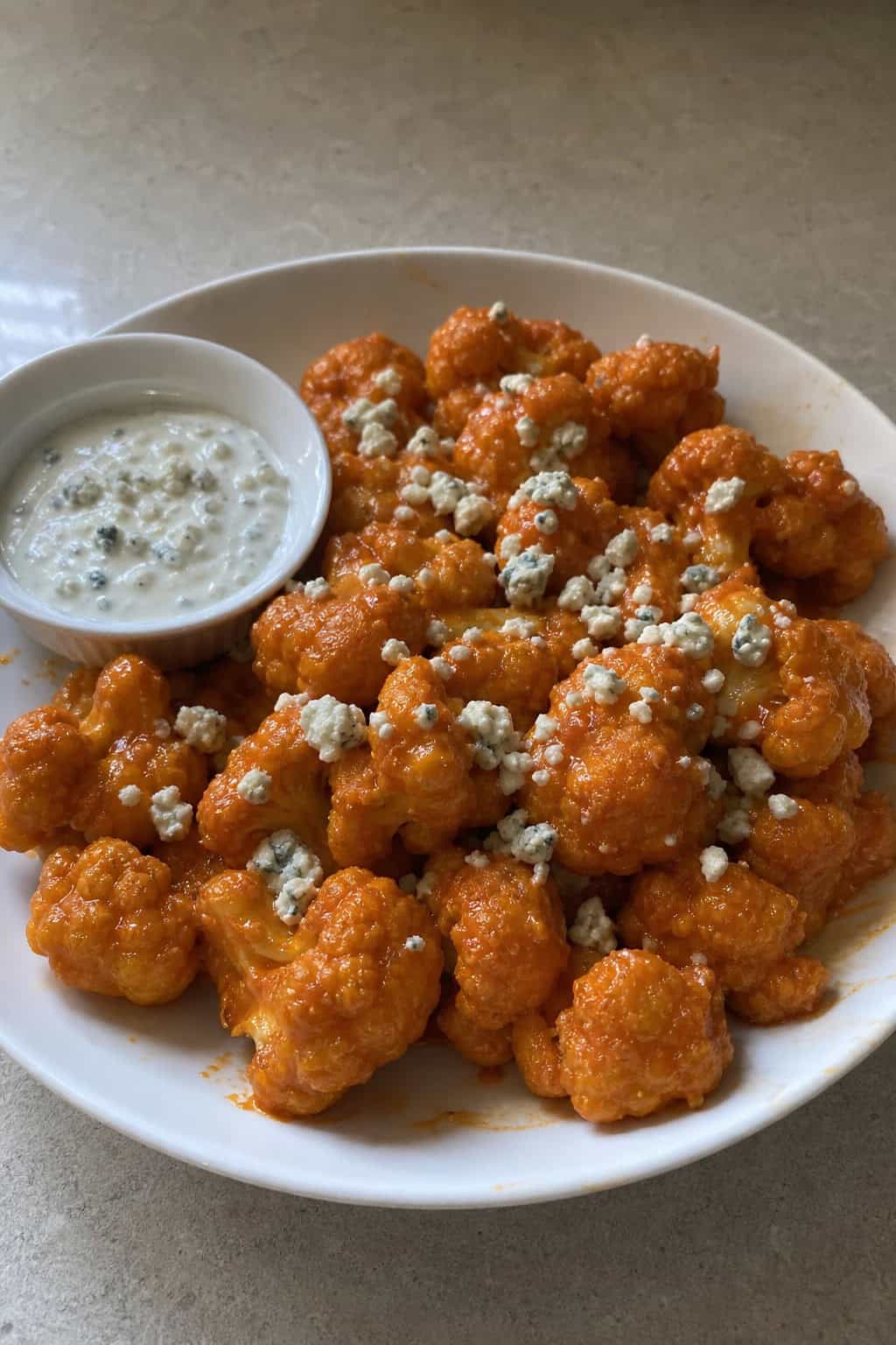 A bowl of buffalo cauliflower bites with blue cheese crumbles and a small cup of blue cheese dressing on a wooden table.